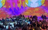 A large group of people sits on cushions in a dark room, watching colourful, abstract animated figures and shapes projected on the walls, creating a vibrant, immersive art experience.