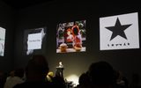 A speaker stands at a lectern in a dark room, addressing an audience. Three large album covers are projected on the wall behind them, including The Next Day and Blackstar by David Bowie.