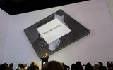 A person stands at a lectern in front of an audience, presenting in a room with a large projection of an album cover titled The Next Day, featuring a black-and-white image partially obscured by a white square.