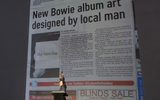 A man stands onstage, pointing at a giant projected newspaper article headlined New Bowie album art designed by local man, as an audience watches. The projection covers the entire wall behind him.