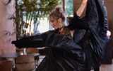 A woman in a dark flowing dress dances gracefully indoors, lifting the fabric as she moves. She has light brown hair in a loose bun and appears focused, with plants and chairs visible in the blurred background.