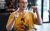 A person with short hair and glasses wears a graphic yellow T-shirt featuring a comic-style figure. They gesture with both fists raised and appear to be speaking or singing. A blurred figure stands in the background.