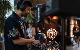 A DJ wearing headphones and a dark shirt with floral embroidery mixes music on a DJ console. A colourful disco ball lights up the scene, with blurred people and indoor plants in the background.