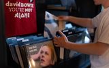 A person browses vinyl records in a shop. In the foreground is a David Bowie album, and a red shirt with the words david BOWIE YOU'RE NOT ALONE hangs above the records.