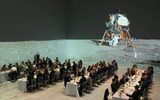 A formal dinner is being held in a large room with a realistic moon surface projection; guests sit at long tables, and an image of a lunar module is displayed in the background, creating a space-themed atmosphere.