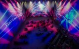 People sit on benches in a dark, immersive room with vibrant purple and blue lights. Large projections on the walls show a person’s face and the words “DON’T LOOK DOWN” repeated multiple times.