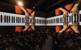 A large audience watches a pianist perform on stage, while giant projections of the pianist’s hands playing the keyboard are displayed on surrounding walls.
