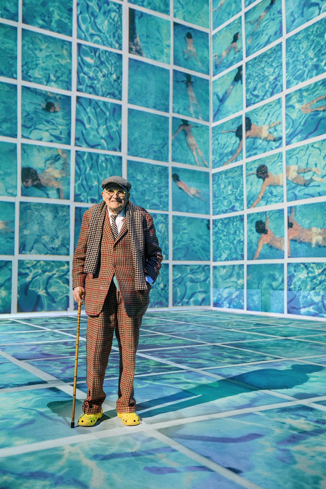 An elderly man in a checked suit, flat cap, and yellow Crocs stands with a walking stick in a room covered floor to ceiling with images of people swimming in a pool. The pattern creates an immersive, surreal effect.
