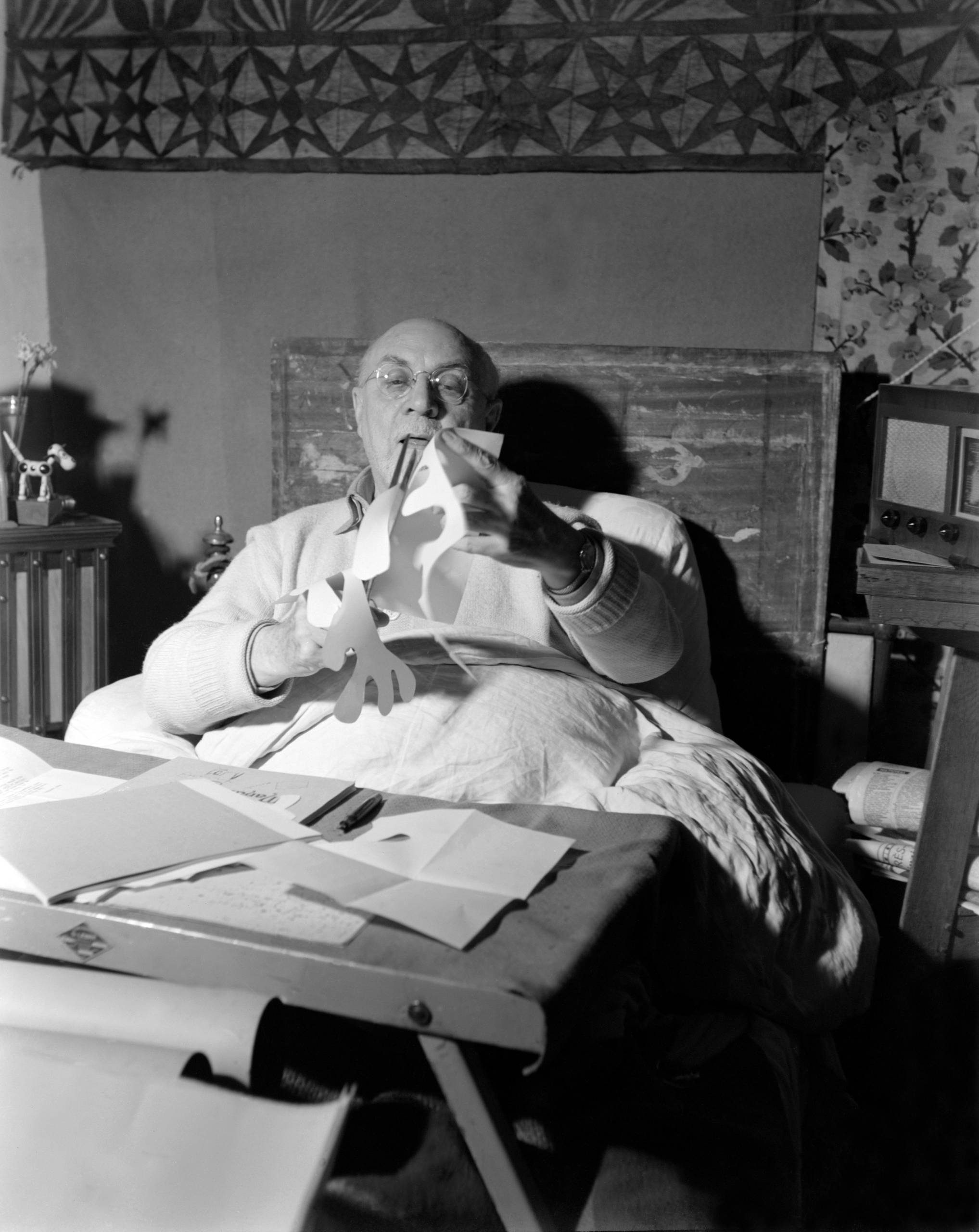 An elderly man sits up in bed, wearing glasses and using scissors to cut paper. A table with papers and art supplies is in front of him. The background features patterned wallpaper and a radio.