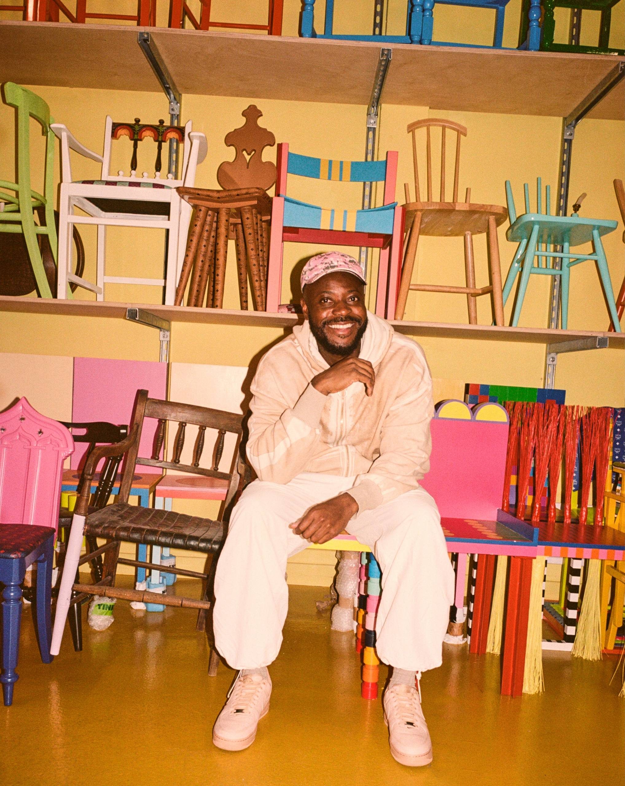 A man in light-coloured clothes and a cap sits smiling in a colourful room filled with various mismatched chairs stacked on yellow shelves behind him.